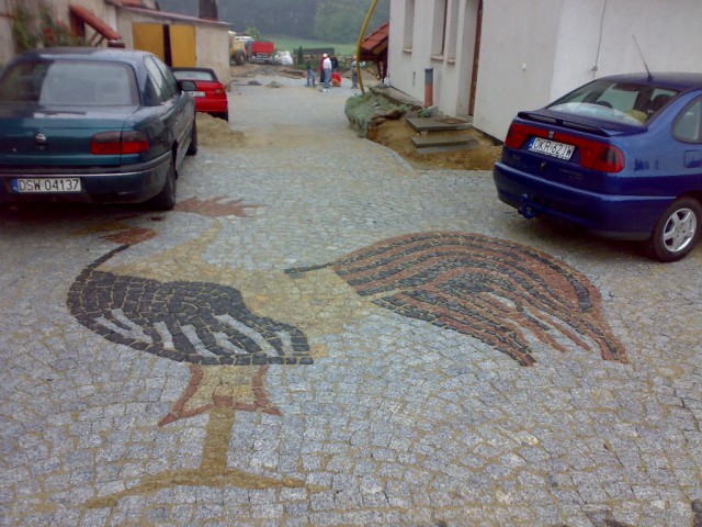 Ułożona z kostki granitowej figura bociana na podjeździe, widoczne dwa samochody osobowe i prace brukarskie w tle.